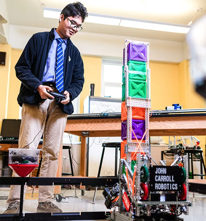Student operating a robot with a remote control in a classroom, next to a tall tower made of colorful plastic cubes labeled 'John Carroll Robotics'.