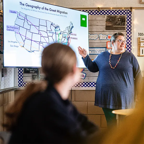 Teacher pointing at a screen displaying a map of the United States titled 'The Geography of the Great Migration' in a classroom with students.