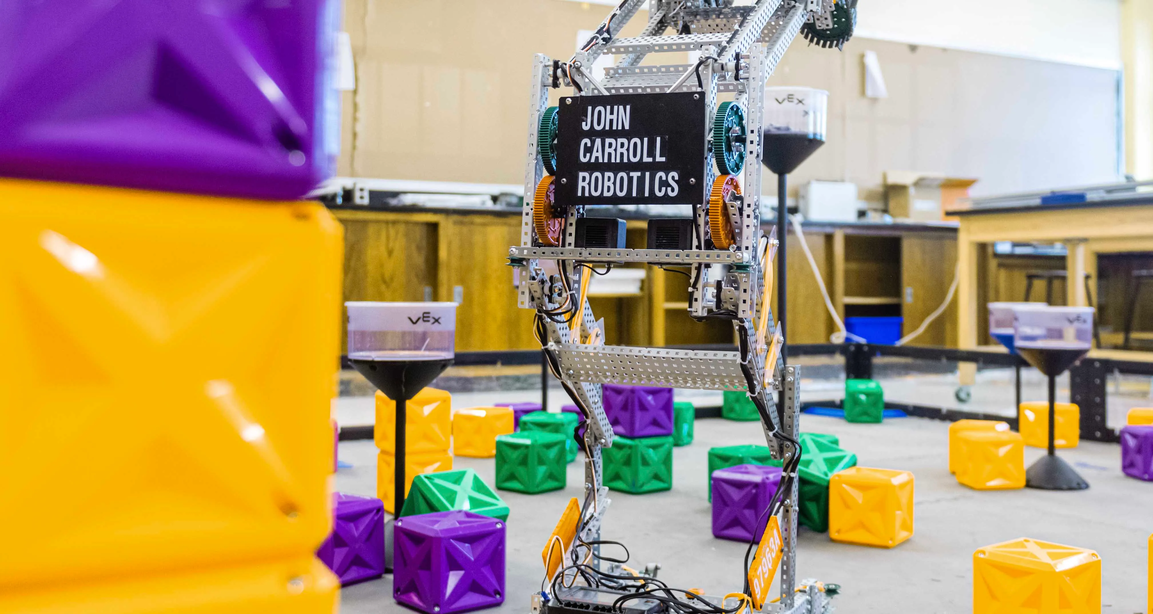 A VEX robotics competition field with a metal robot labeled 'John Carroll Robotics' surrounded by colorful cube game elements in yellow, green, and purple.