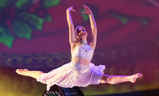 Ballet dancer in white costume performing a grand jeté with arms raised on stage.