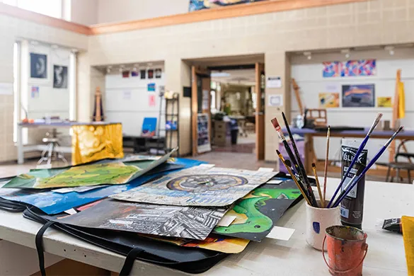 Art classroom table with various colorful paintings, paintbrushes in containers, and paint tubes, with an open doorway and artwork displayed on walls in the background.
