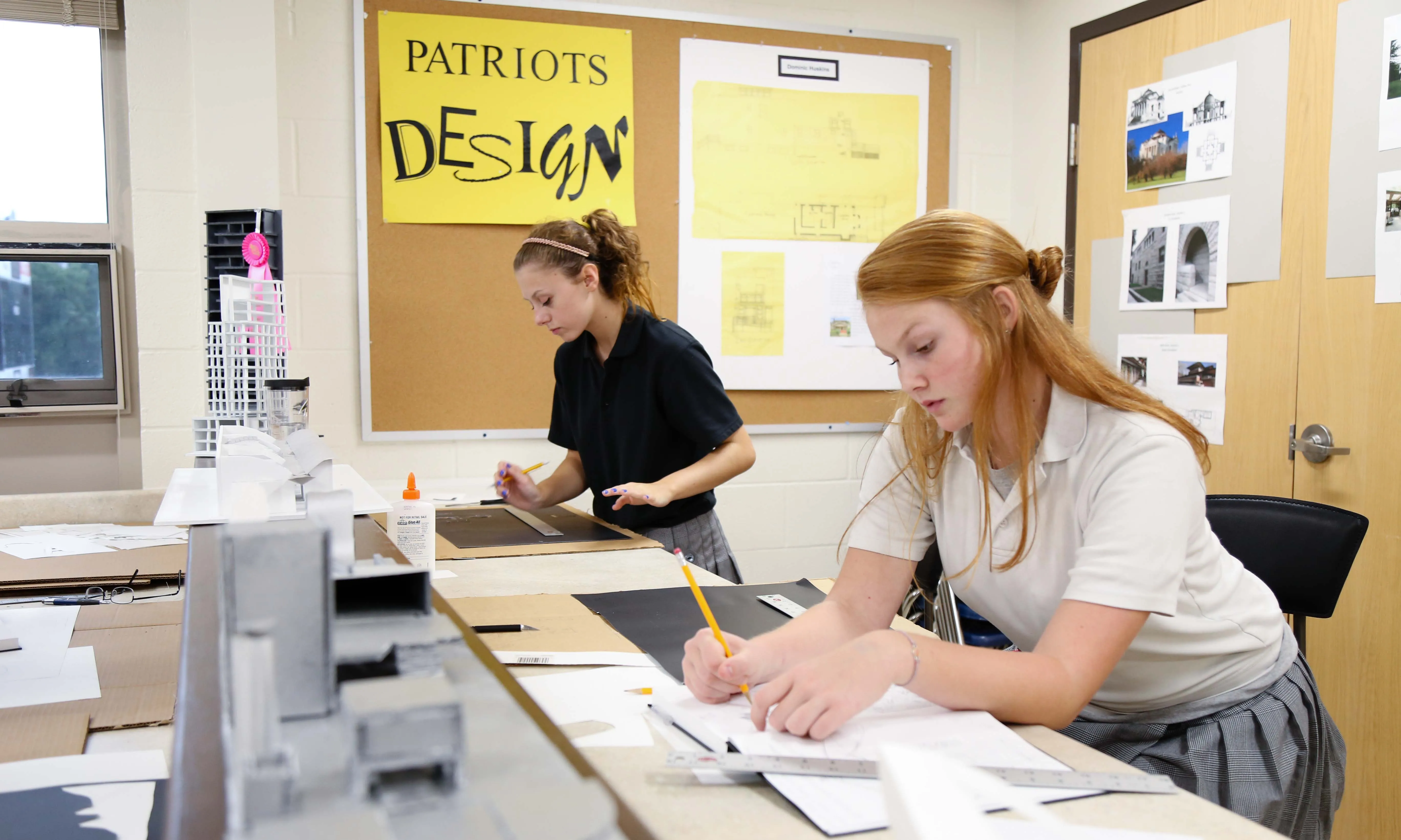 Two female students working on architectural projects at a classroom table with design models and drawings.