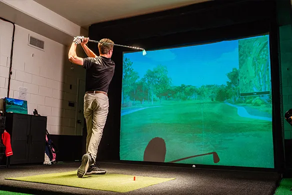 Man swinging a golf club indoors in front of a large screen showing a virtual golf course.