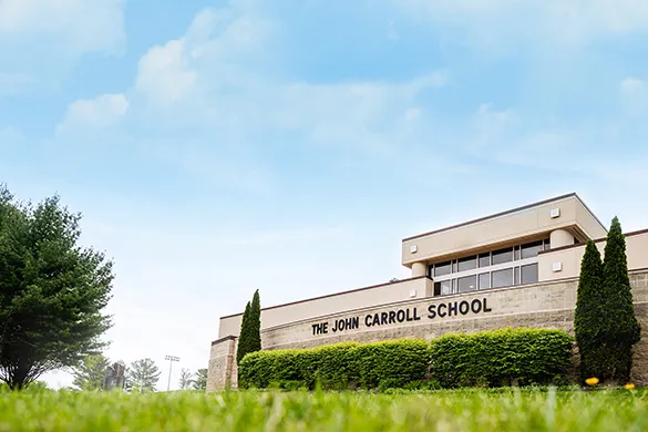 Exterior view of The John Carroll School building under a partly cloudy sky with green grass and trees in the foreground.