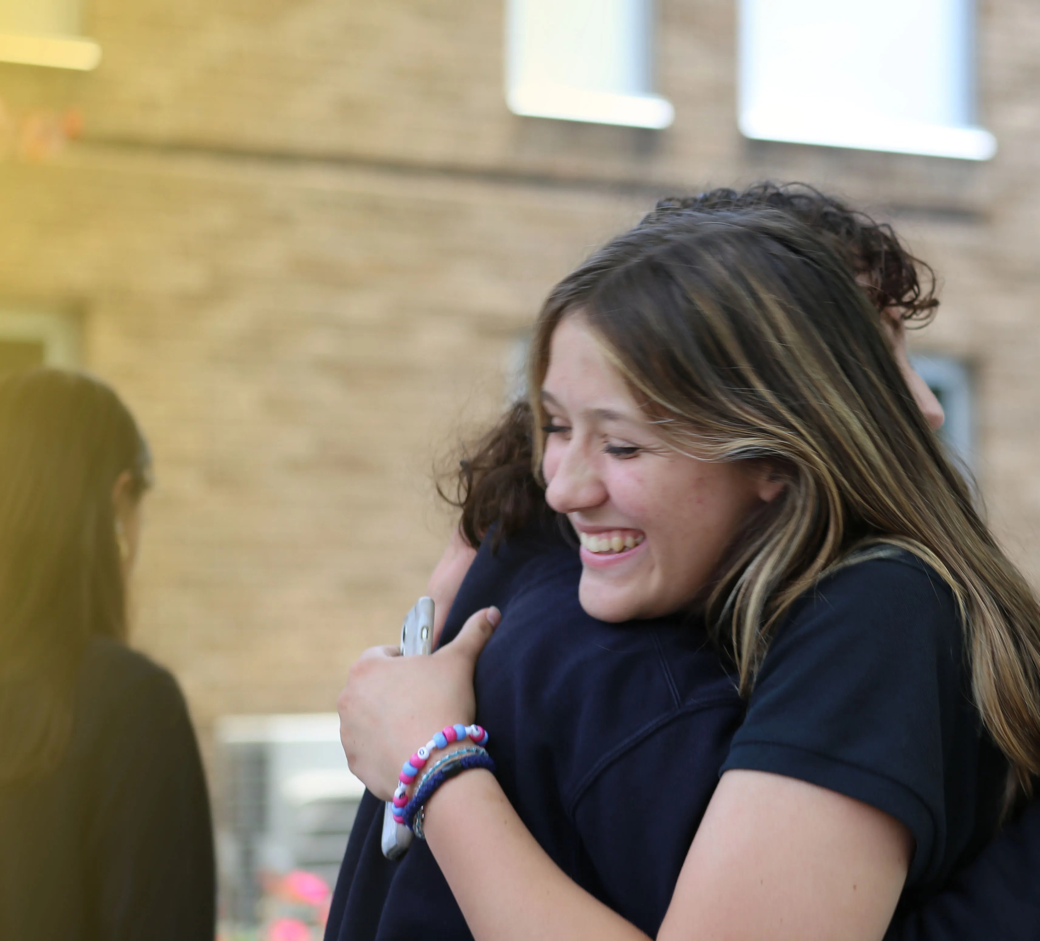 Two young people hugging joyfully outdoors with one smiling broadly, holding a phone and wearing colorful beaded bracelets.