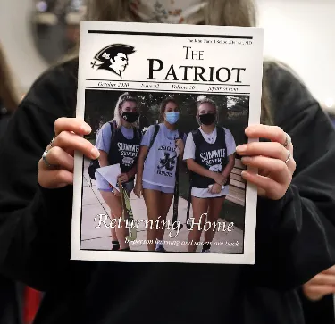 Person holding a copy of The Patriot newspaper featuring three masked students in summer school shirts under the headline 'Returning Home.'
