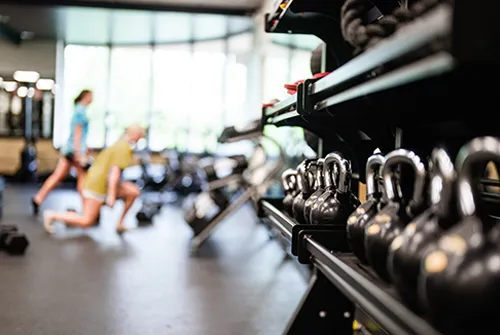 Row of kettlebells on rack in a gym with two people exercising in the background.