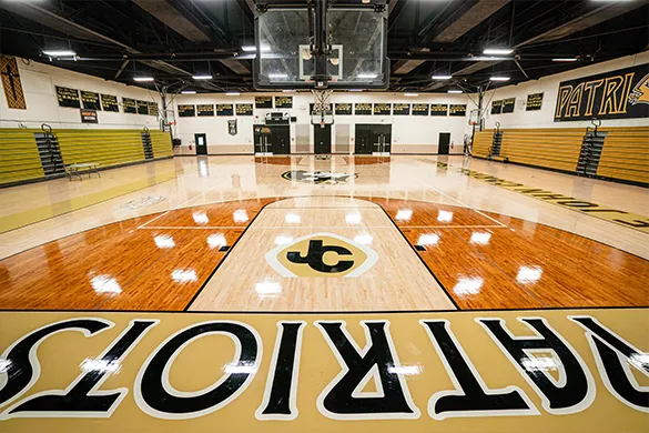 Empty indoor basketball court with 'Patriots' text on the floor and bleachers on both sides.