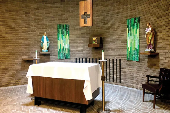 Interior of a small chapel with brick walls, an altar covered with a white cloth, two statues on either side, pillar candles, and a wooden crucifix on the wall.