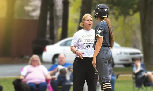 Softball coach in white Patriots t-shirt talks to player in black uniform standing on field.
