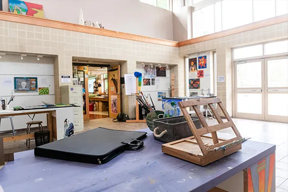 Art studio workspace with easel, paint supplies, and portfolio case on a table near large windows and an open doorway.