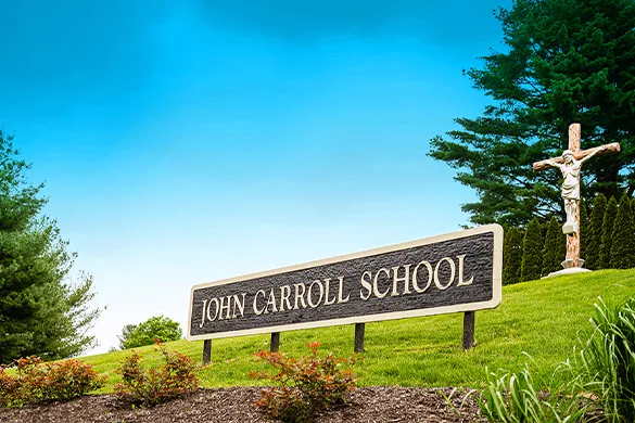 Sign reading 'John Carroll School' on a grassy hill with trees and a crucifix statue in the background under a clear blue sky.