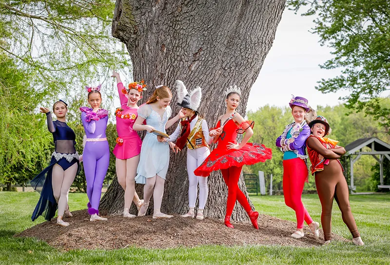 Group of eight children in colorful ballet costumes posing near a large tree outdoors on grass.