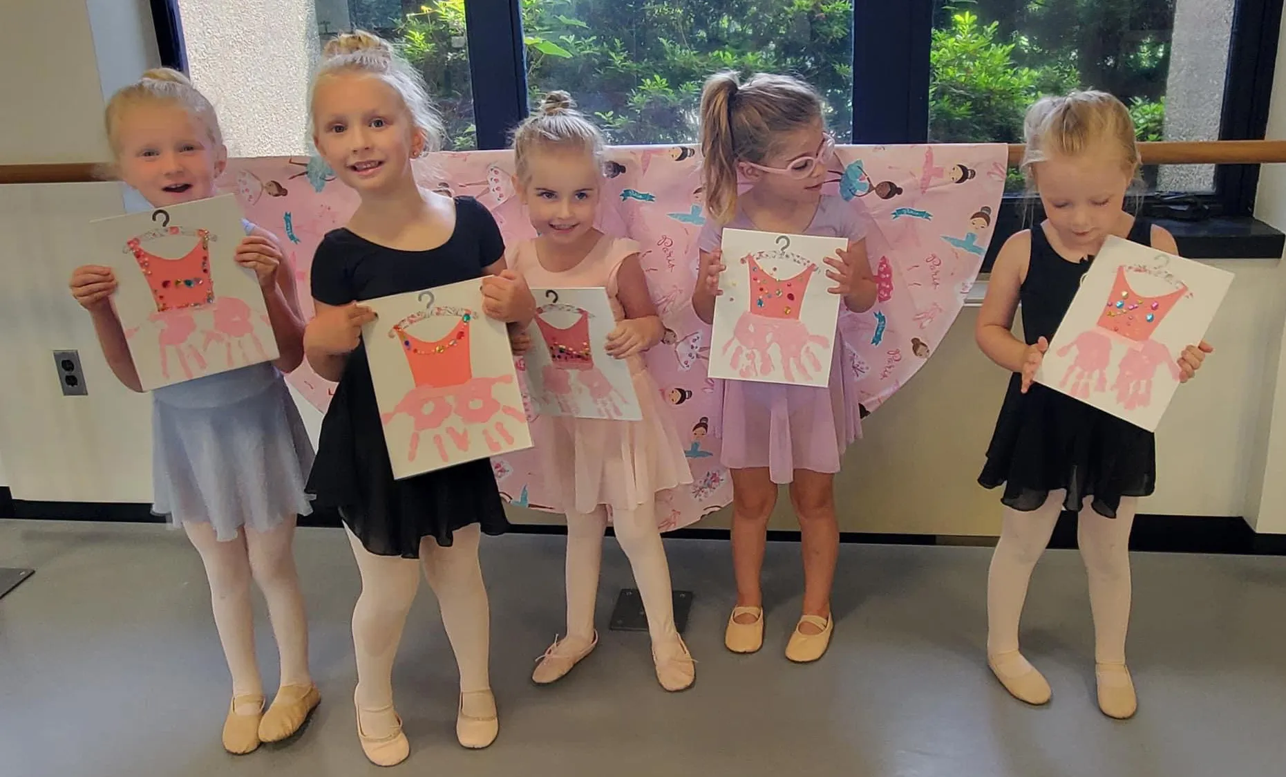 Five young girls in ballet outfits holding artwork of pink dresses decorated with handprints and gems.