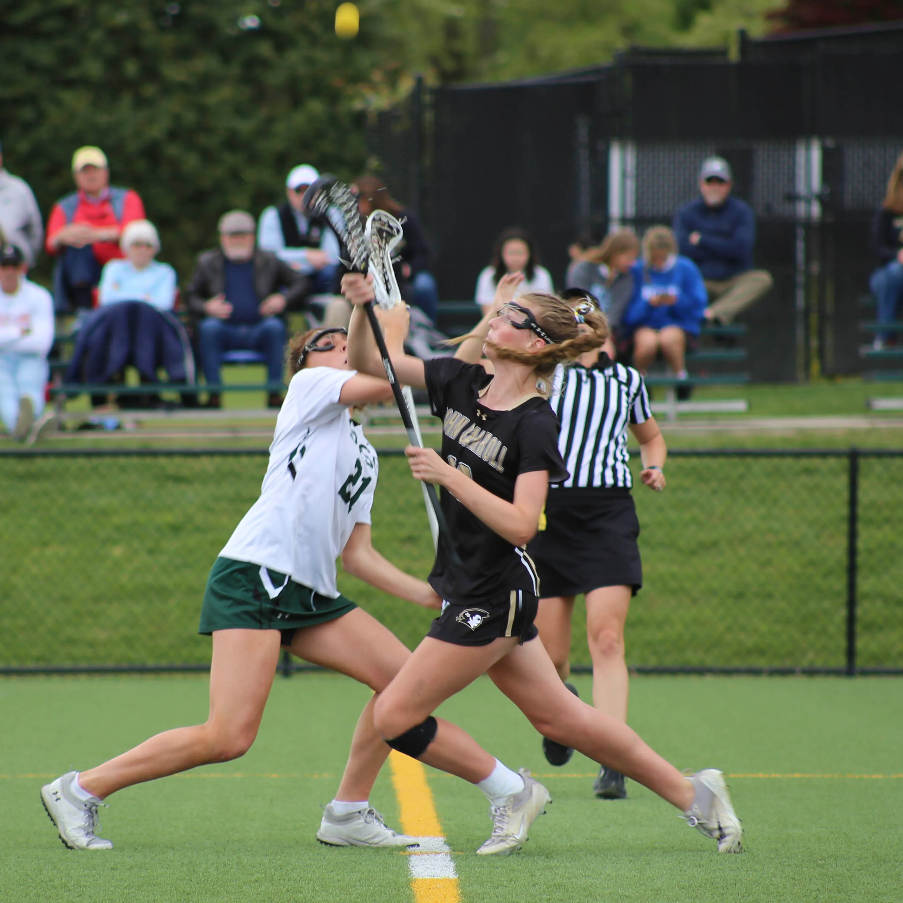 Two female lacrosse players in mid-action competing for the ball on a green field with an official and spectators in the background.