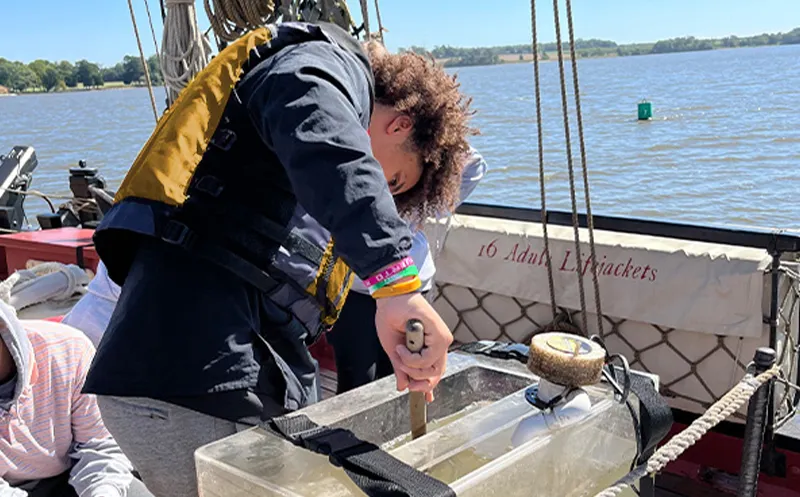 Person wearing a life jacket using a tool on a clear rectangular container on a boat with water and land in the background.