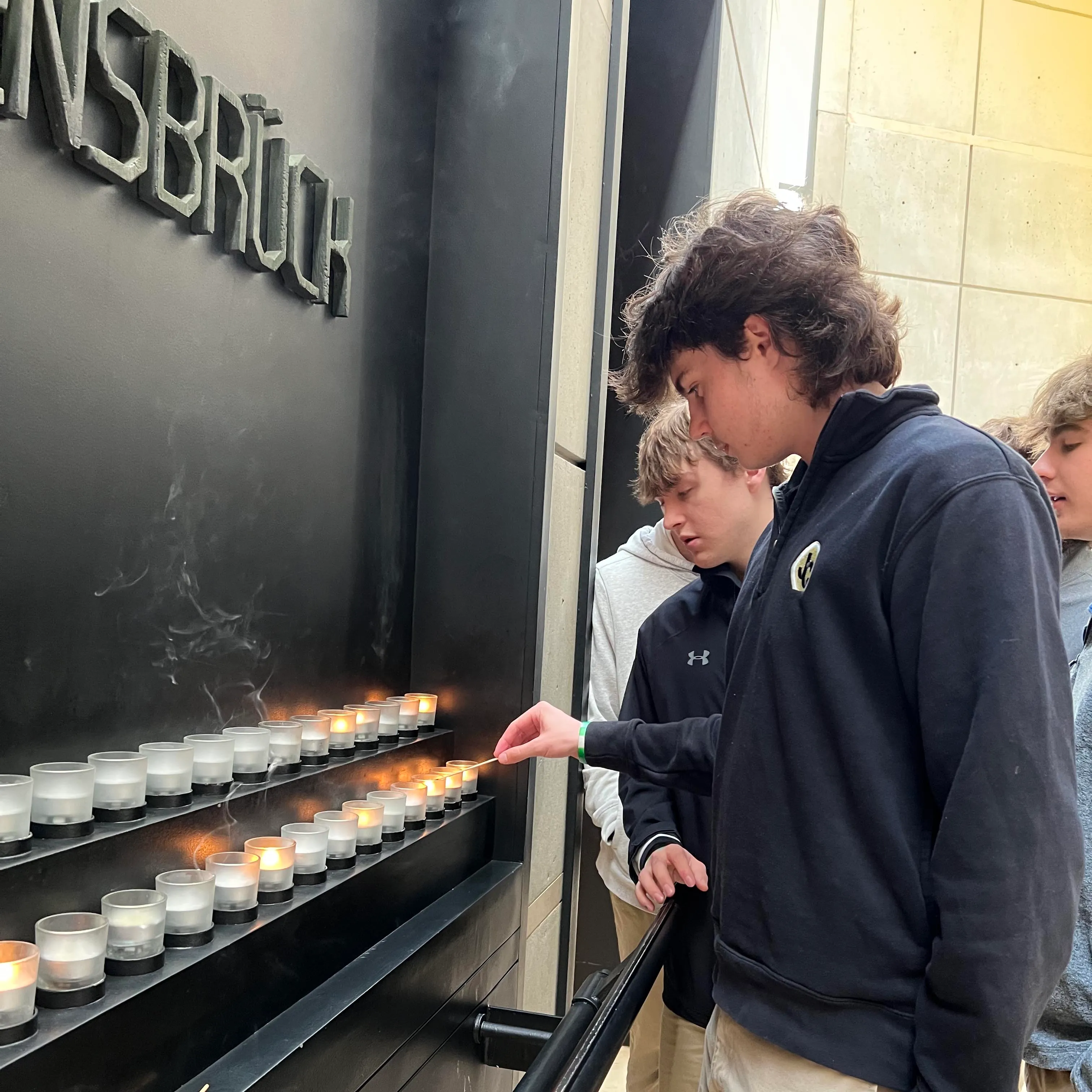 Three young men lighting candles at a memorial wall with the word 'AUSBRÜCK' visible above.