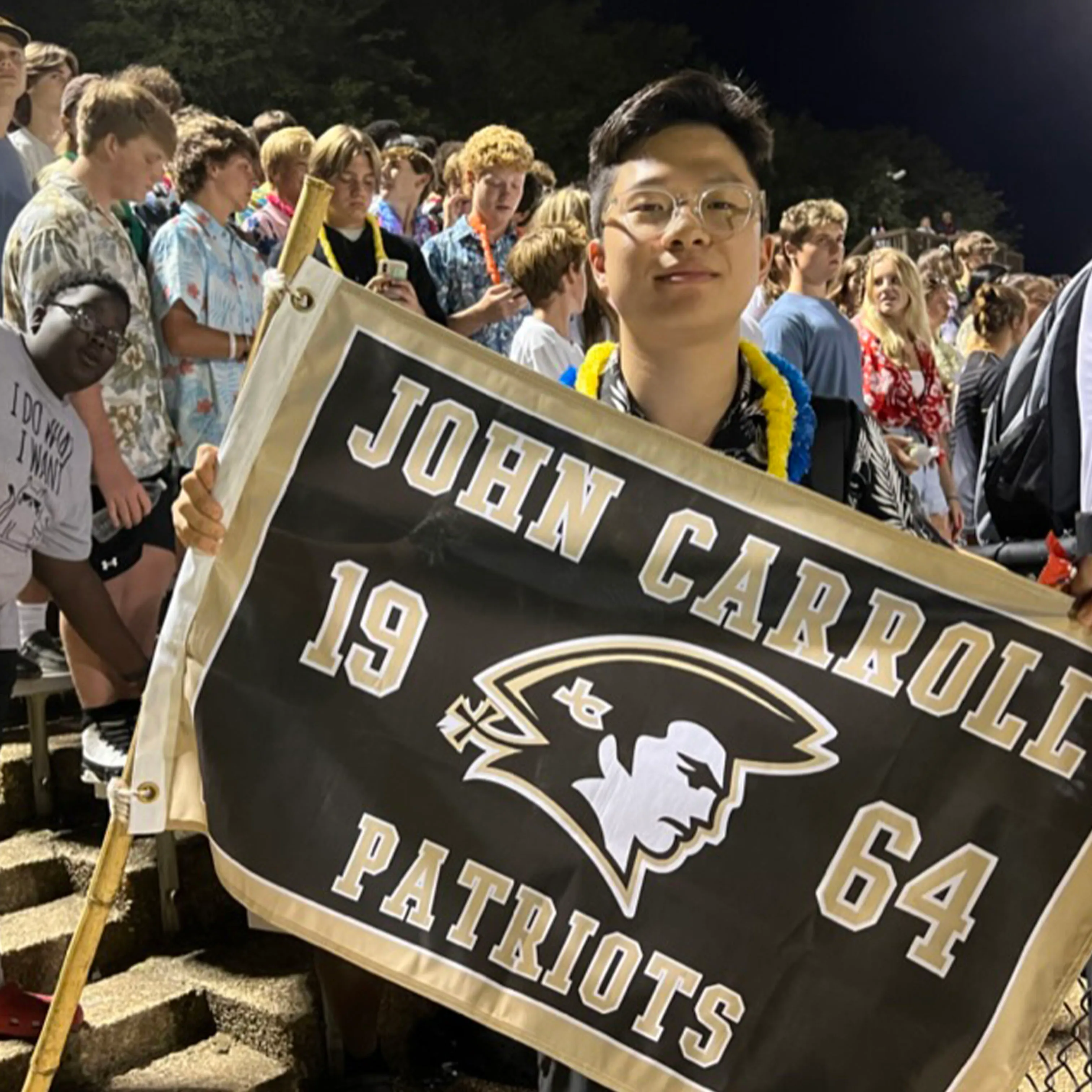 A young man wearing glasses holds a John Carroll Patriots flag with the years 1964 and a patriot logo, standing among a crowd at a night event.