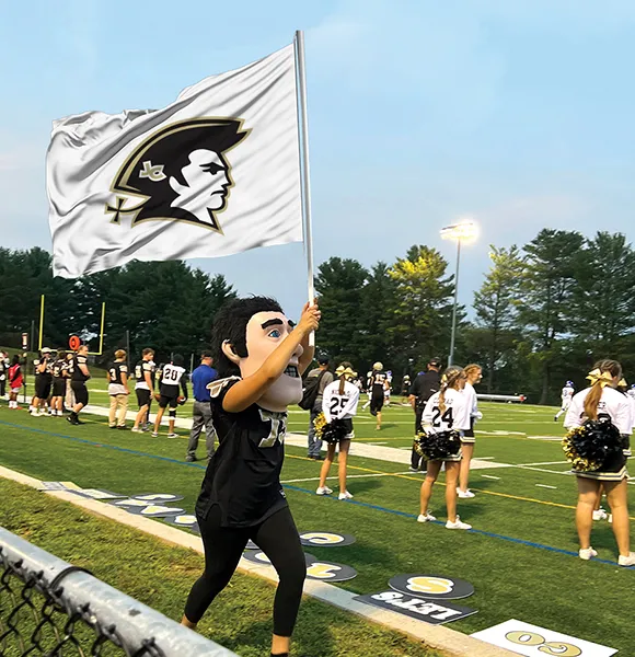 Mascot with large head costume waving a white flag with a black and gold profile logo on a football field with cheerleaders and players in the background.