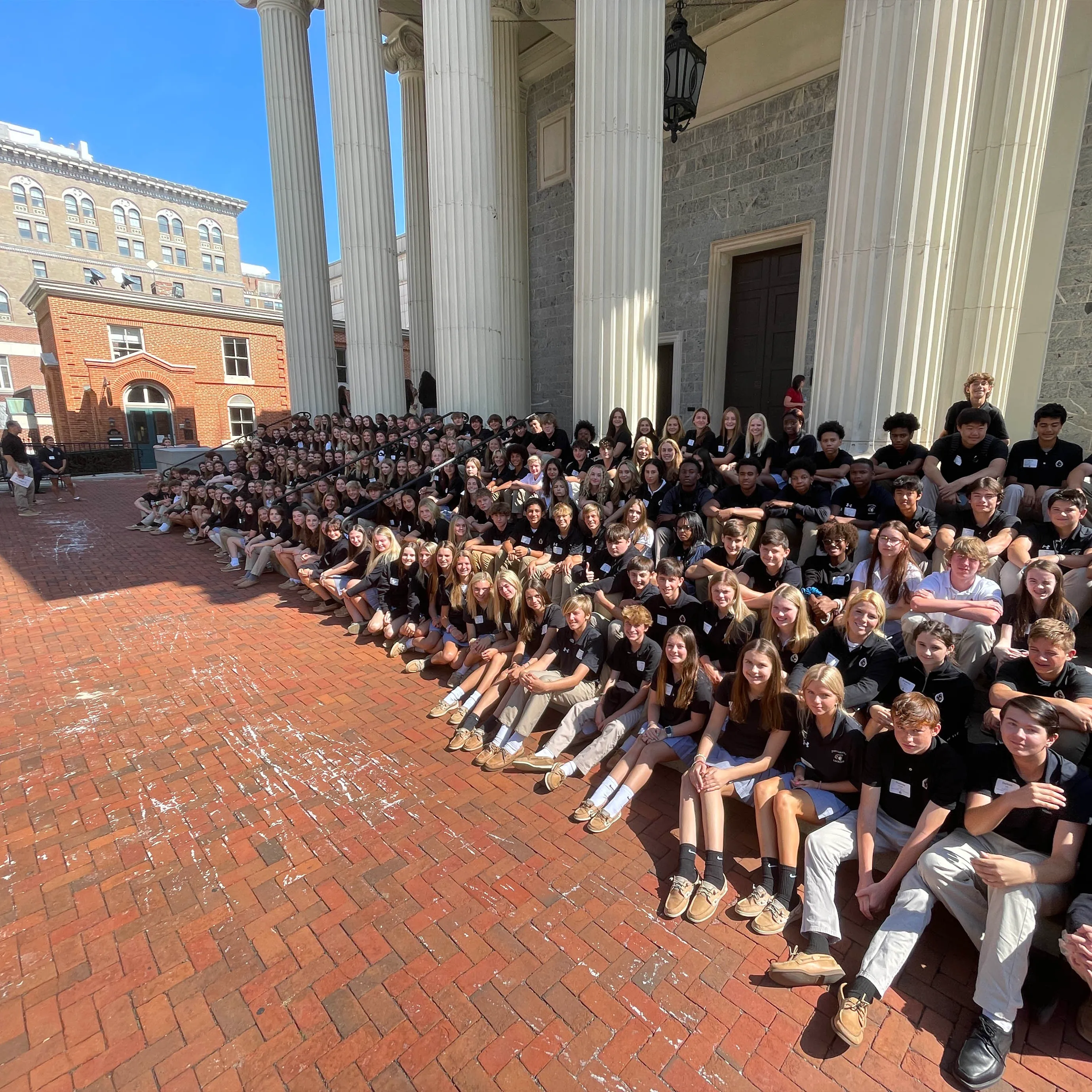 Large group of students sitting on steps and pavement in front of a building with tall white columns on a sunny day.