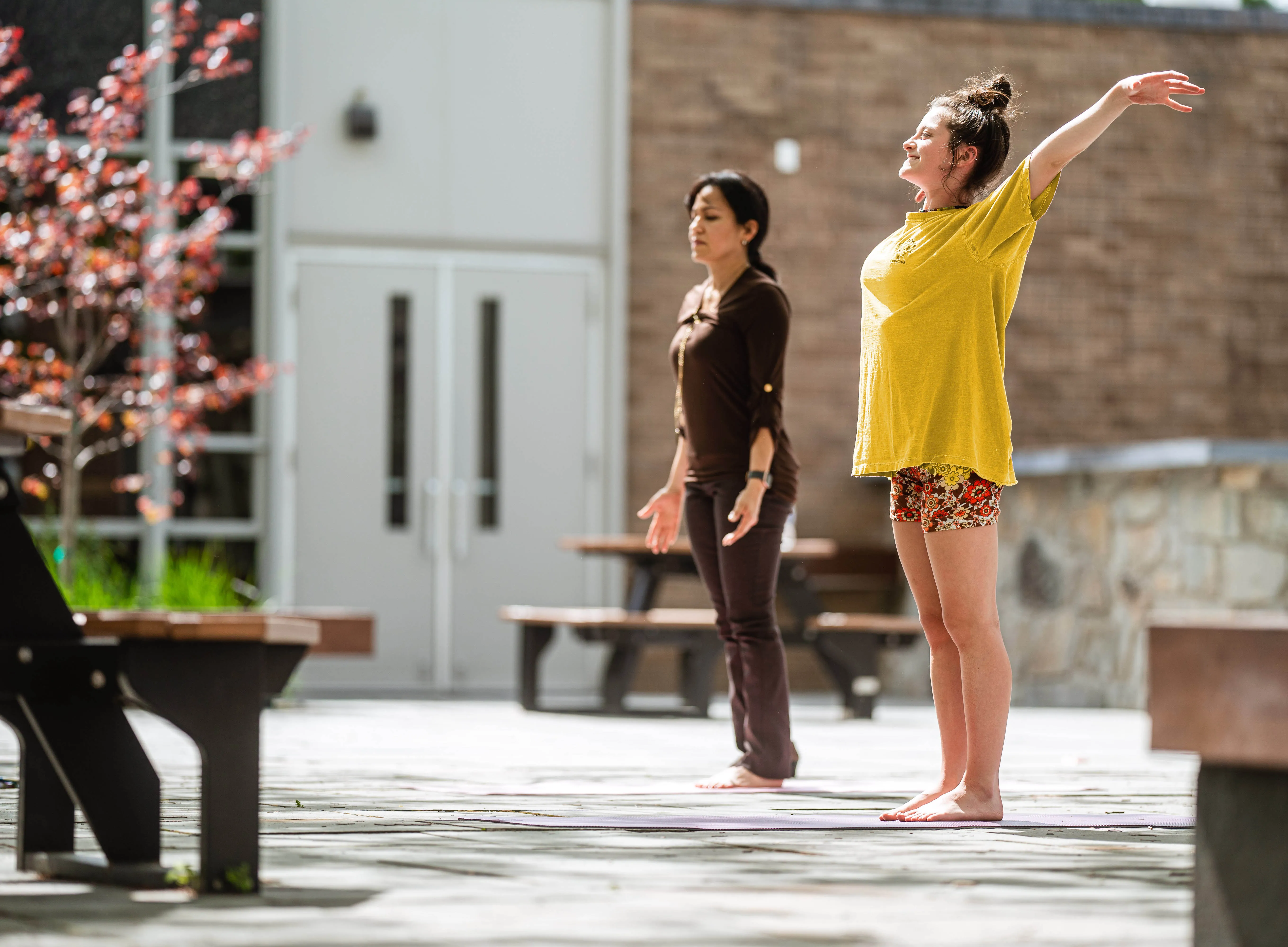 Two women practicing yoga outdoors in a sunlit courtyard, one stretching arms upwards and the other standing with arms by her sides.