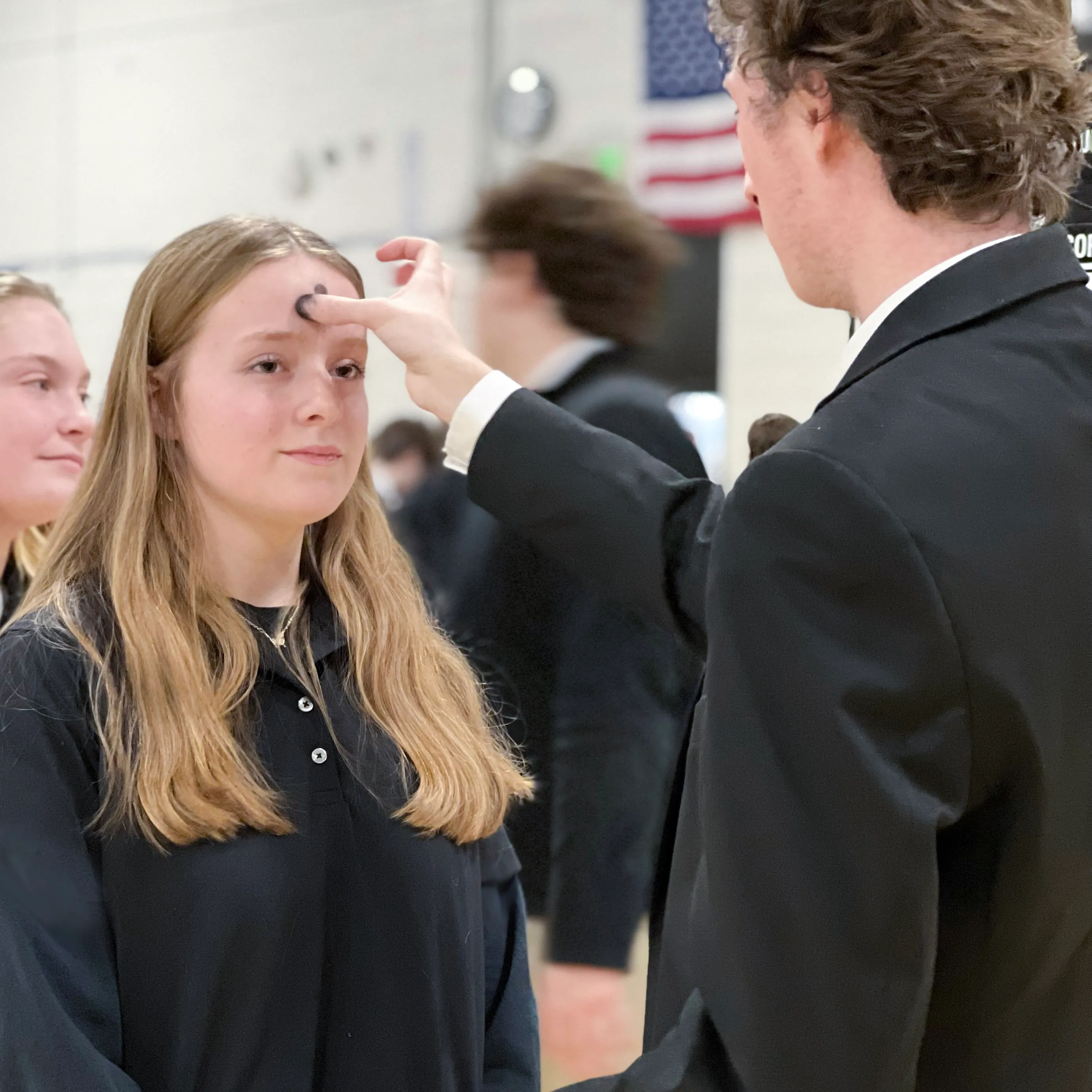 A young man in a suit marks a black dot on a young woman's forehead, who is standing with closed lips indoors.