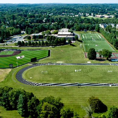 Aerial view of a school campus featuring a running track surrounding a soccer field, a football field, and campus buildings, with trees and residential areas in the background.