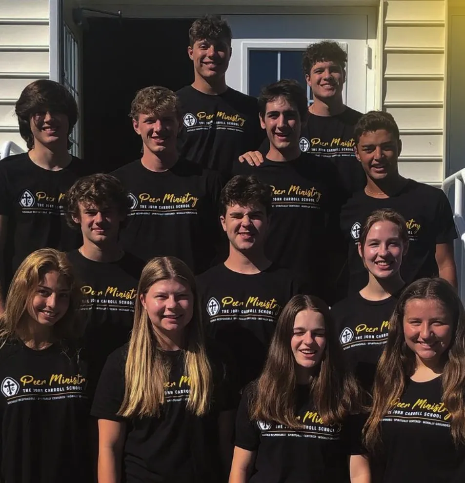 Group of smiling teenagers wearing matching black Peer Ministry T-shirts standing outside a building entrance.
