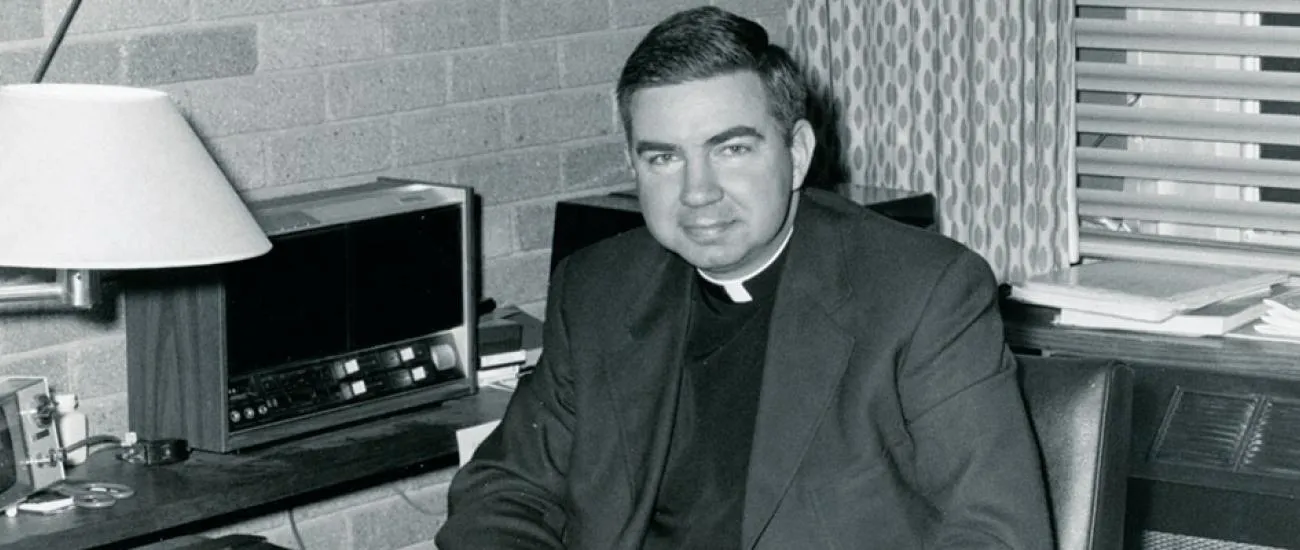 Black and white photo of a man wearing a clerical collar and suit jacket sitting in an office with a lamp, vintage radio equipment, and patterned curtains.