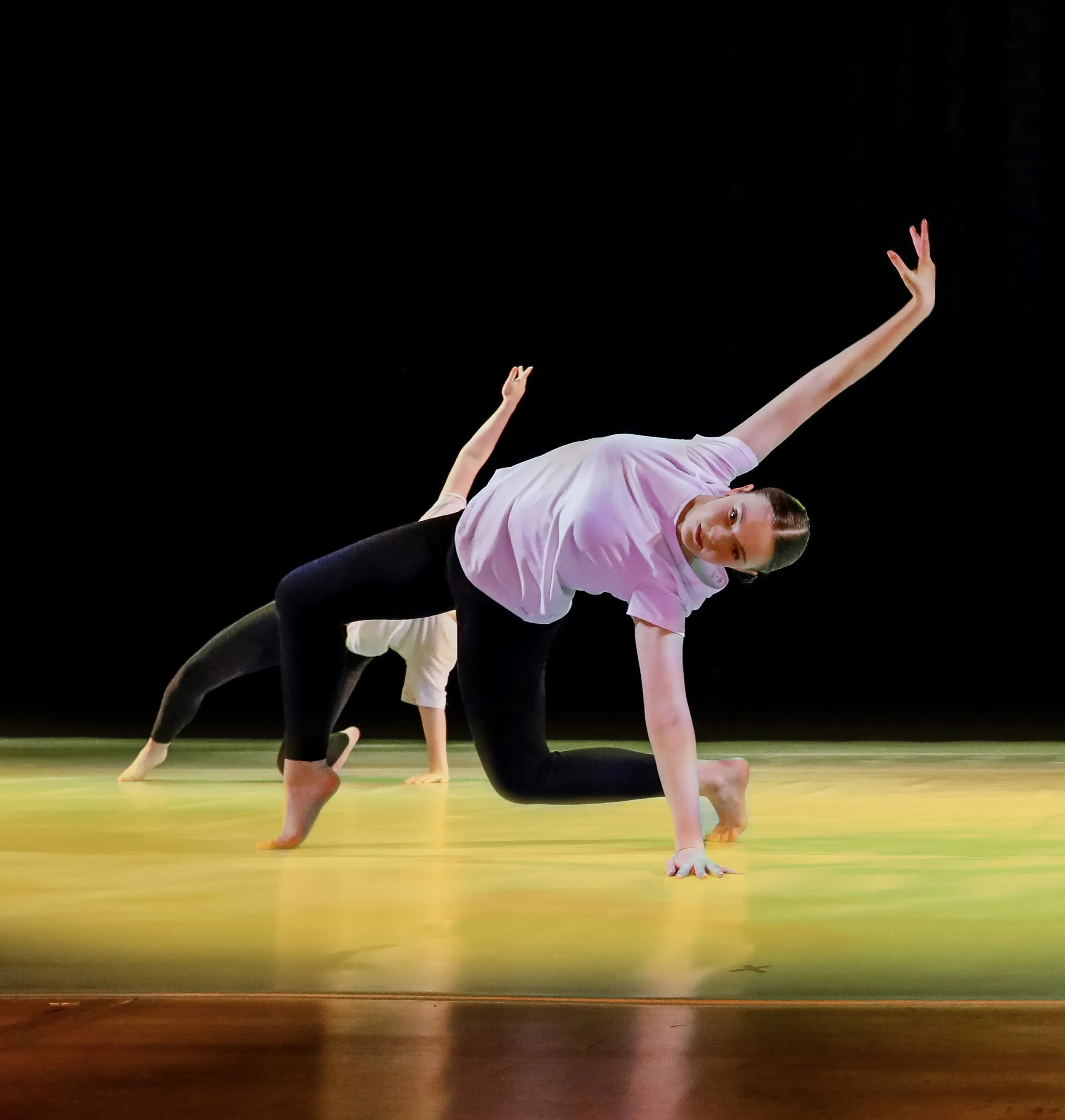 Two dancers performing a contemporary floor pose with one hand and one foot on the ground and the other arm extended upwards on a lit stage.