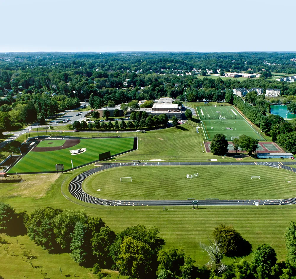 Aerial view of a sports complex featuring a baseball field, a soccer field, tennis courts, and a running track surrounded by trees and greenery.