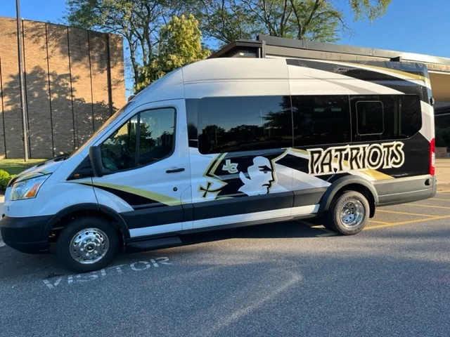 Black and white mini bus with gold accents featuring a patriot's profile logo and the word 'PATRIOTS' on the side, parked in a lot.