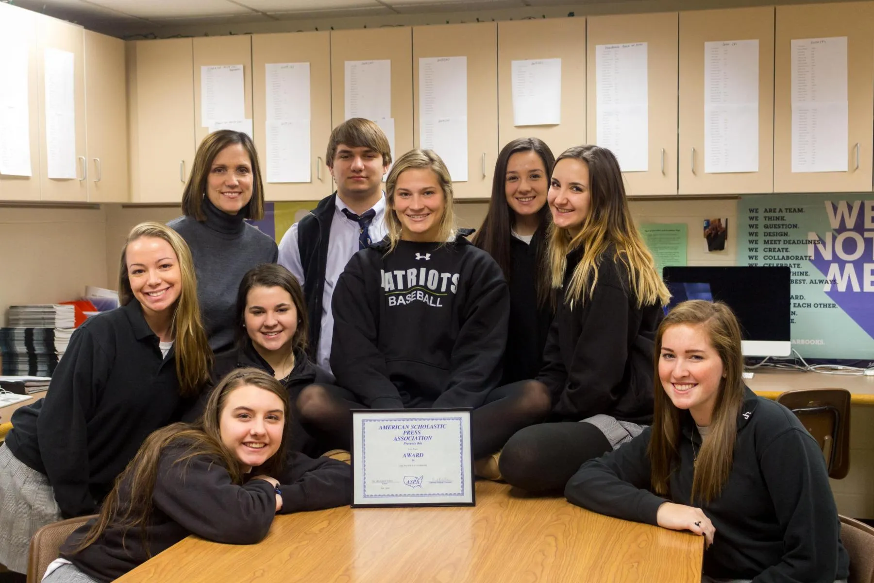 Group of nine smiling students and one teacher gathered around a table in a classroom with an American Scholastic Press Association award displayed on the table.