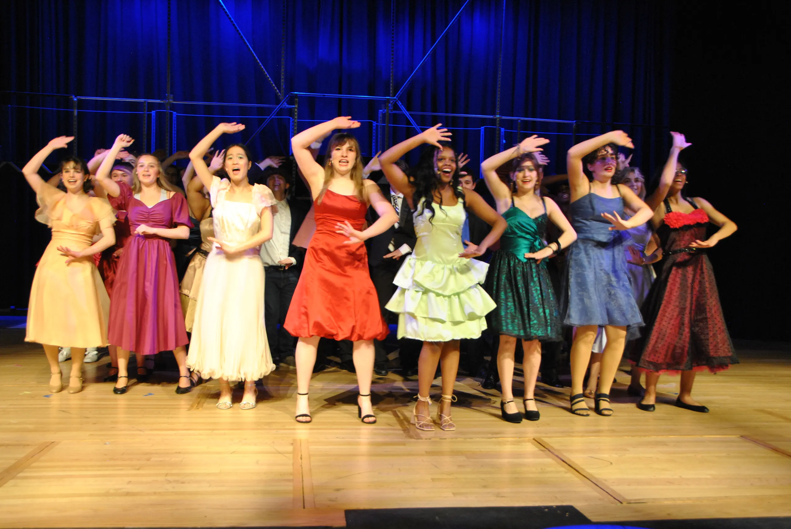 Group of performers in colorful dresses dancing on stage with hands raised, under blue stage lighting.
