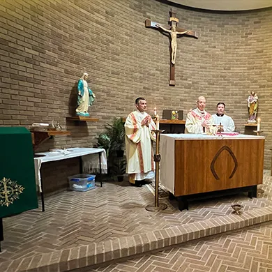 Three priests in white and gold vestments standing behind an altar in a brick chapel with religious statues and a crucifix on the wall.