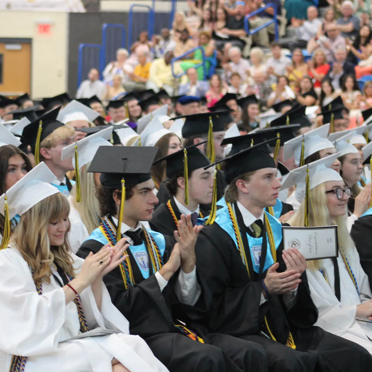 High school graduates in white and black caps and gowns sitting and clapping during a graduation ceremony with audience in the background.