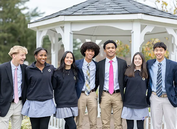 Group of seven diverse high school students in uniforms standing together outdoors in front of a white gazebo, smiling.