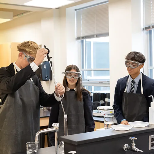 Three students wearing aprons and safety goggles conduct a chemistry experiment in a bright science lab.
