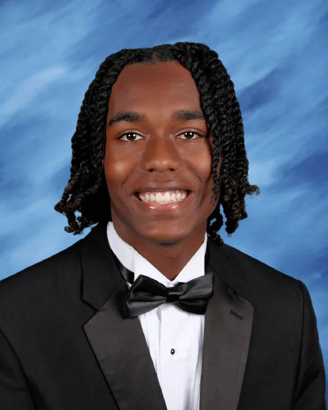 Smiling young man with braided hair wearing a black tuxedo and bow tie against a blue background.