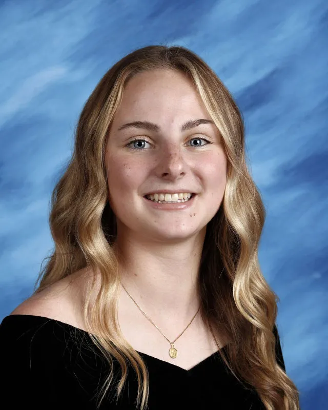 Young woman with long wavy blonde hair wearing a black off-the-shoulder top and a gold necklace, smiling against a blue marbled background.