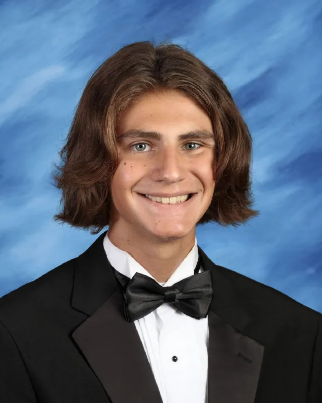 Young man with shoulder-length brown hair wearing a black tuxedo, white shirt, and black bow tie, smiling against a blue backdrop.