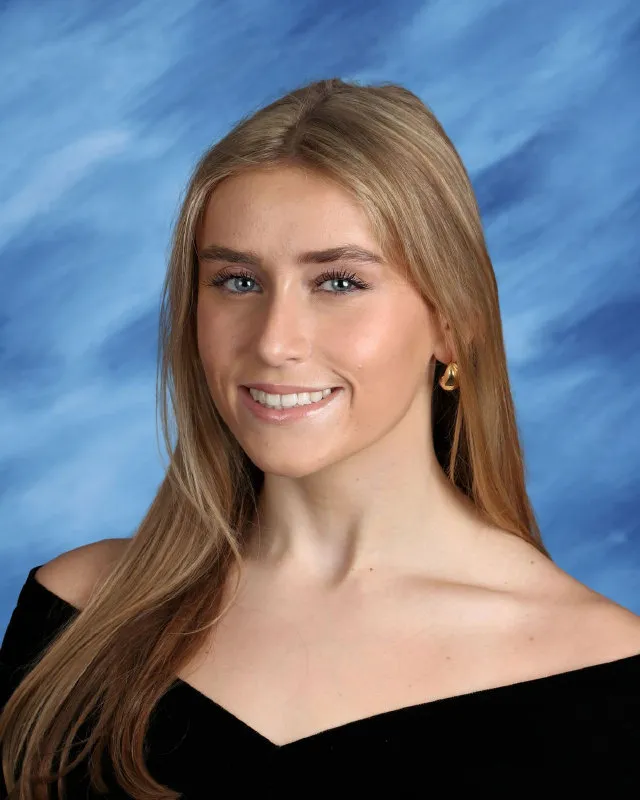 Young woman with long blonde hair wearing a black off-the-shoulder top and gold earrings against a blue studio background.