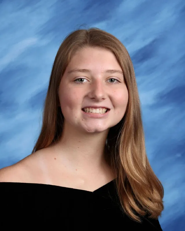 Young woman with long straight blonde hair smiling against a blue cloudy backdrop wearing a black off-shoulder top.
