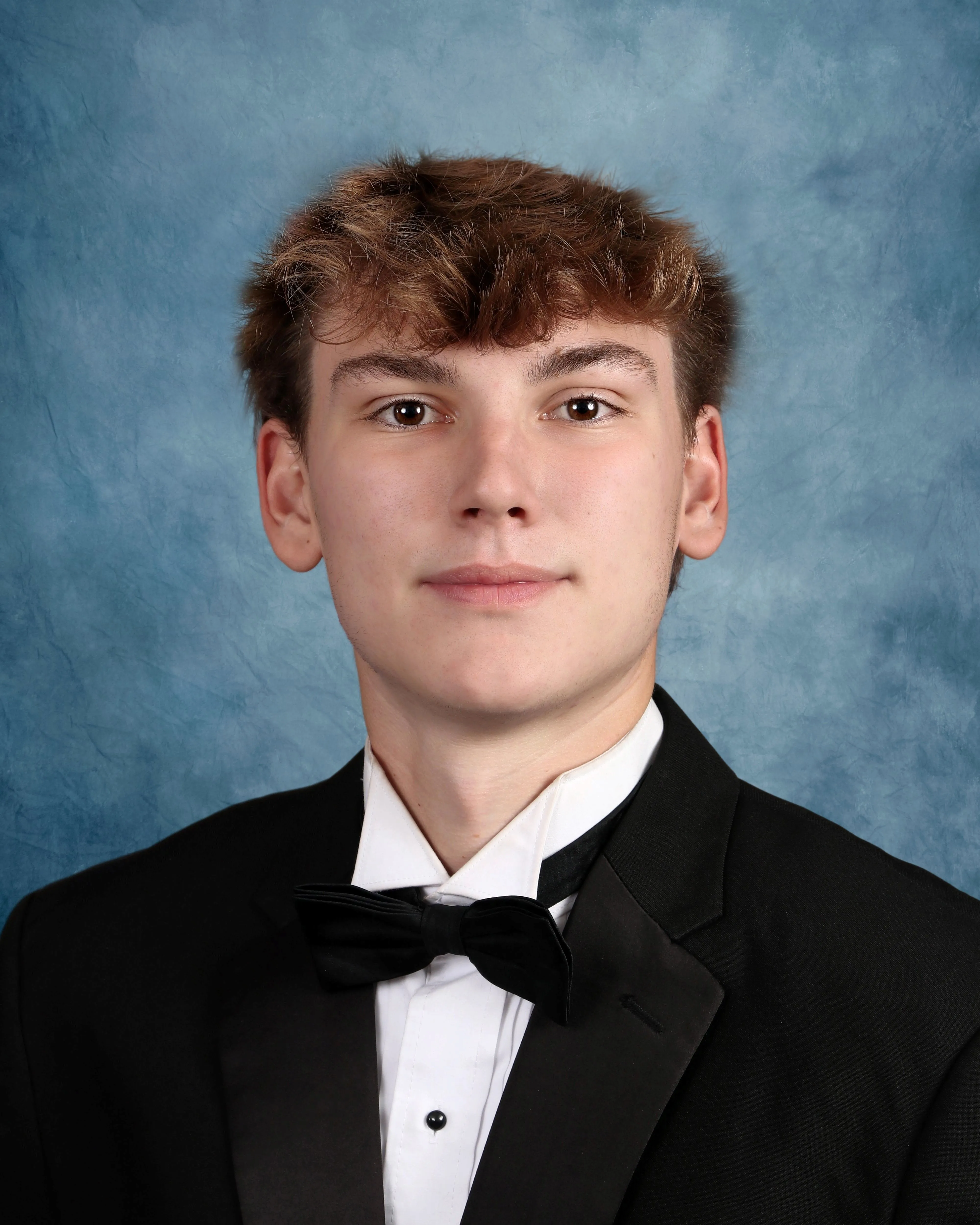 Young man with light brown hair wearing a black tuxedo jacket, white shirt, and black bow tie against a blue textured backdrop.
