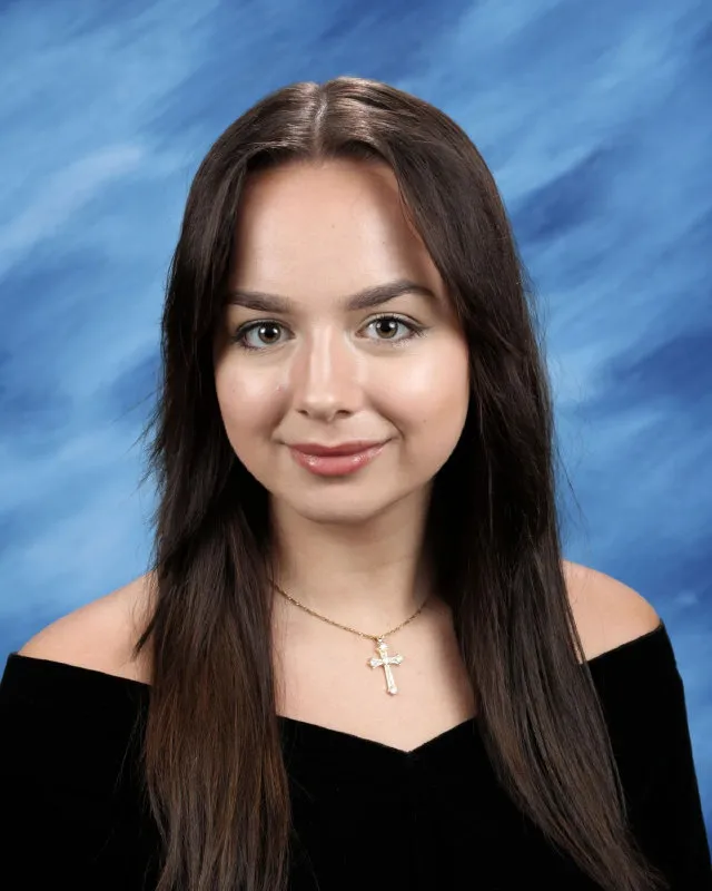Young woman with long brown hair wearing a black off-shoulder top and a gold cross necklace, smiling against a blue background.