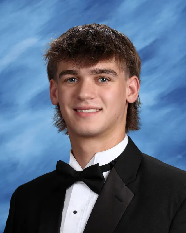Young man with brown hair wearing a black tuxedo and bow tie against a blue textured background.