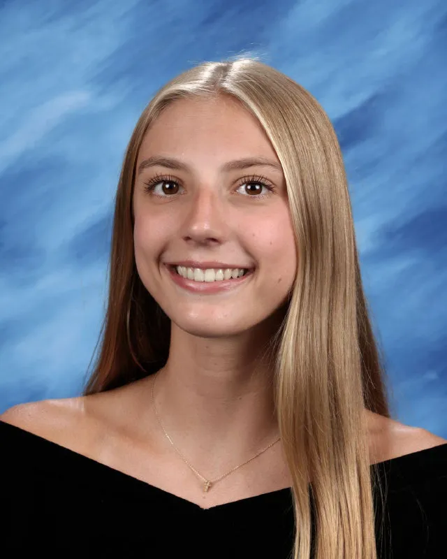 Smiling young woman with long blonde hair wearing a black off-shoulder top and a delicate necklace against a blue studio background.