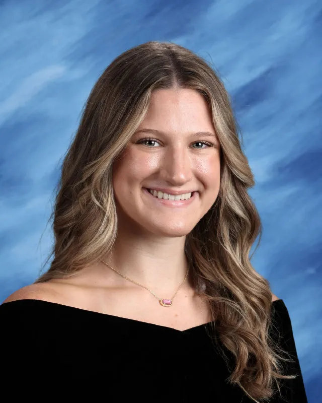 Smiling young woman with long wavy blonde hair wearing a black off-shoulder top and a pink pendant necklace against a blue backdrop.