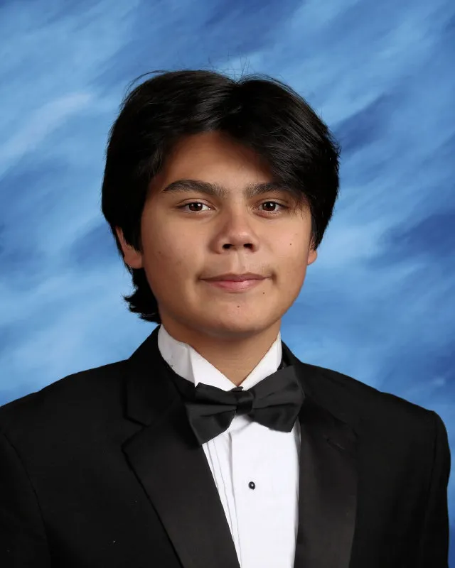 Portrait of a young man with black hair wearing a black tuxedo, white shirt, and black bow tie against a blue backdrop.