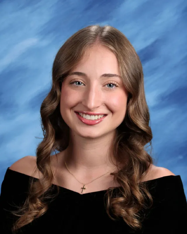 Young woman with long curly light brown hair wearing an off-shoulder black top and a gold cross necklace, smiling against a blue studio background.